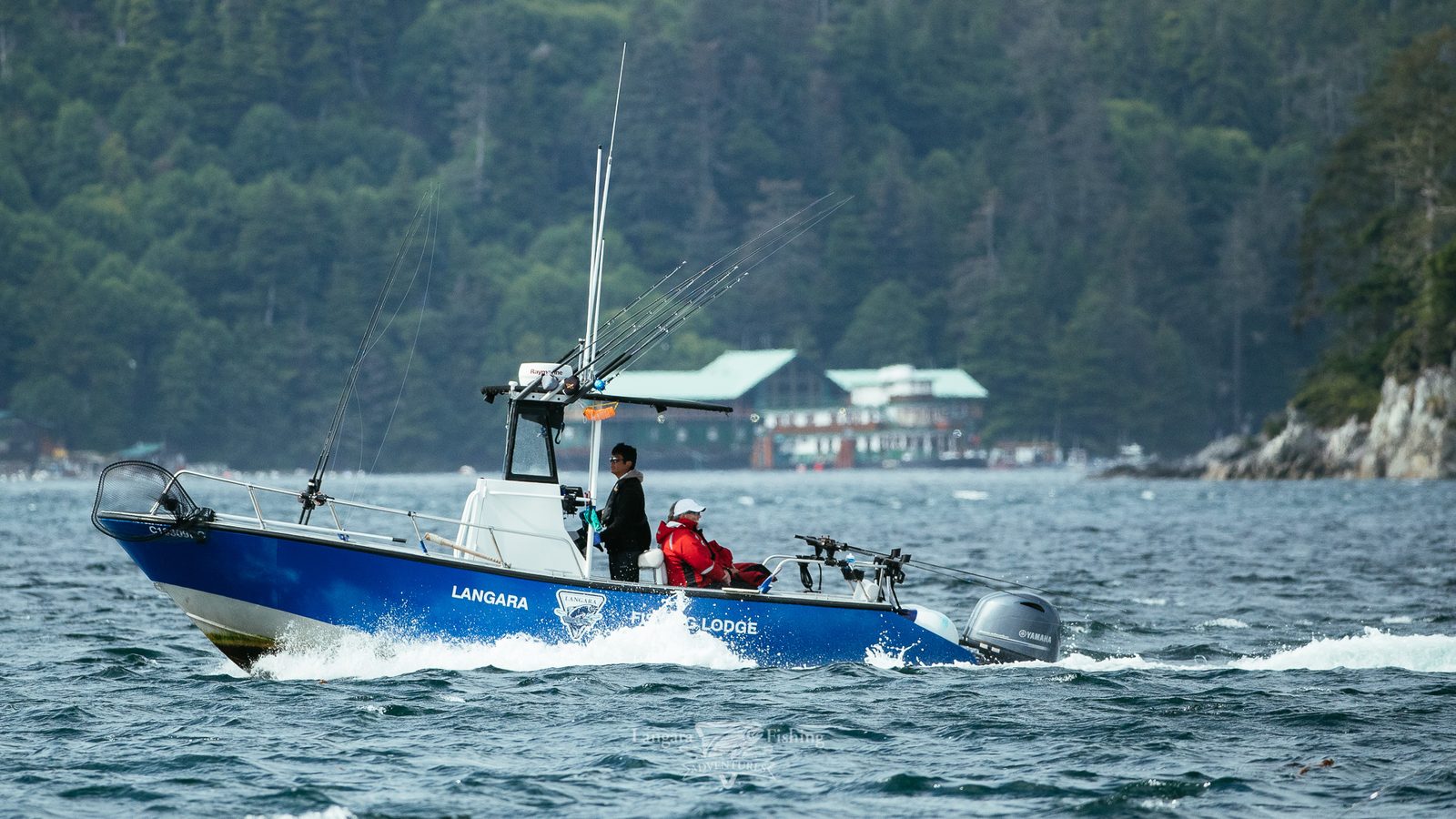 A blue Langara Fishing Lodge boat running across the water, the lodge behind it.
