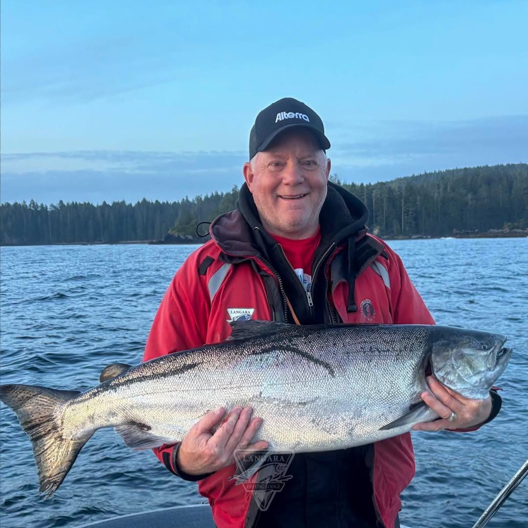 An angler in a red jacket holding a big chinook salmon across his chest, forest and ocean behind.