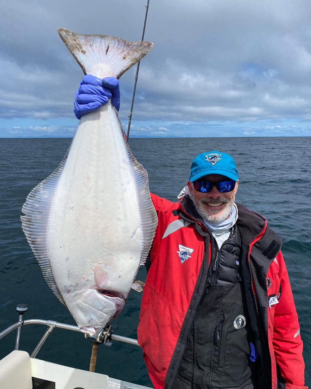 A grinning angler in a red Langara jacket holding up a big halibut on the boat.